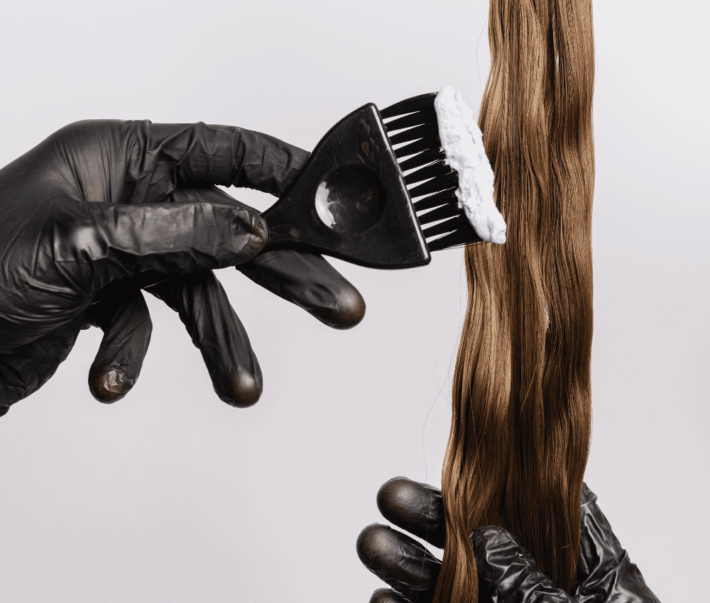 Gloved hand applying hair dye to long brown hair with a brush.